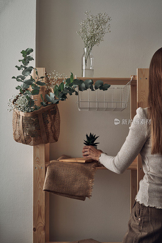 The girl decorates a wooden rack with an eco-bag with eucalyptus branches, flowers, houseplants. Eco-materials, storage. Folk.